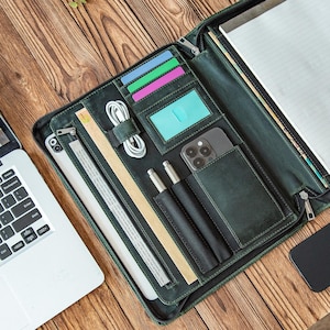 Può includere: Cartella in pelle verde scuro con più scomparti, tra cui porta carte, portapenne e una tasca per il telefono. La cartella è aperta, rivelando un blocco per appunti, un tablet e un laptop. Sono visibili anche un cavo di ricarica bianco e un righello.