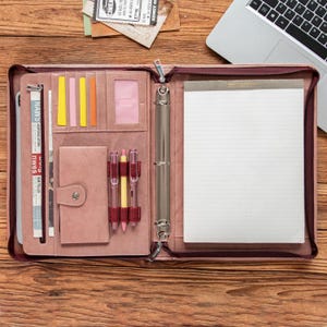 May include: A pink leatherette zipped binder, open to show a notepad, pen holders, and pockets. The binder contains a notepad, pens, and a small notebook. The word "NAWS" is visible on a newspaper clipping. A laptop is partially visible in the background.