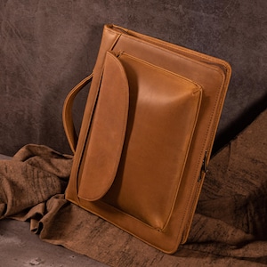 May include: A tan leather portfolio with a handle and a front pocket. The portfolio has a zip closure and is made of smooth, textured leather. The design is simple and functional, suitable for carrying documents and other essentials.