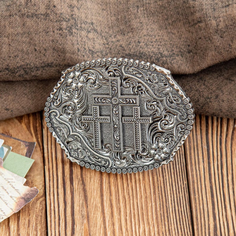 May include: Silver-toned oval belt buckle featuring three crosses at the center, surrounded by floral and scrollwork designs. The buckle has a detailed, textured appearance and a beaded border. The background is a wooden surface.