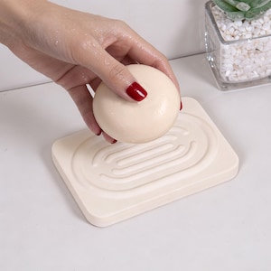 May include: A round, light beige bar of soap is held above a matching rectangular soap dish. The dish has a recessed, patterned design. The background is a white surface with a small potted plant visible in the upper right corner.