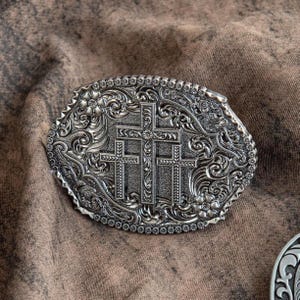 May include: Silver-toned oval belt buckle featuring three crosses in the center, surrounded by floral and scrollwork designs. The buckle has a detailed, textured appearance and a beaded border. A religious-themed accessory.