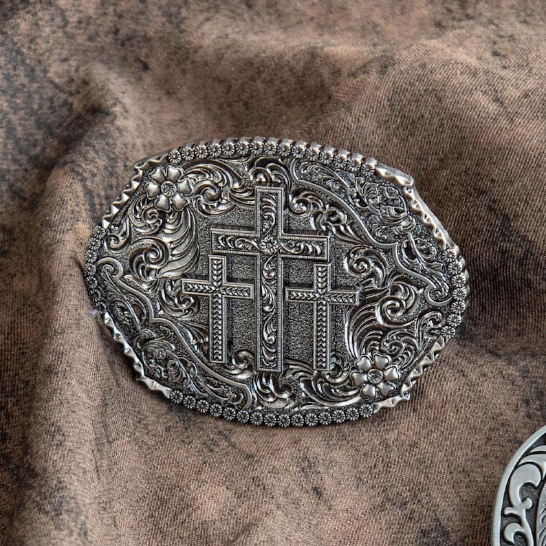 May include: Silver-toned oval belt buckle featuring three crosses in the center, surrounded by floral and scrollwork designs. The buckle has a detailed, textured appearance and a beaded border. A religious-themed accessory.