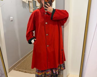 Puede incluir: Un abrigo rojo de manga larga con cuello y puños en verde oscuro, cierre frontal de botones y dobladillo recto. Se lleva sobre una falda de cuadros multicolor, combinada con botines color canela y un sombrero fedora del mismo tono.