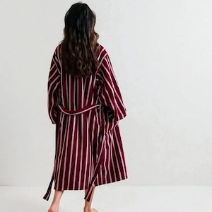 Pode incluir: Um roupão listrado bordô e branco com cinto a condizer. O roupão vai até ao joelho e tem mangas largas. A pessoa está a usar o roupão e está de costas para a câmara.
