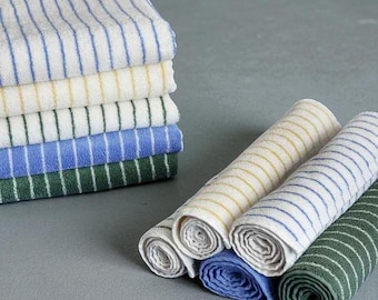 Op de afbeelding: Een stapel gestreepte washandjes in verschillende kleuren: wit met blauwe strepen, crème met gele strepen, wit met groene strepen, blauw met witte strepen en groen met witte strepen. Gerolde washandjes in bijpassende kleuren zijn ook aanwezig.