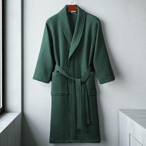 May include: A dark green waffle-weave robe hanging on a wooden hanger. The robe has a shawl collar, two front pockets, and a matching belt. The robe is made of a textured fabric.