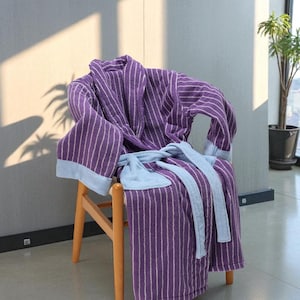 May include: A purple striped bathrobe with light blue trim and belt is draped over a wooden chair. The robe is made of a soft, absorbent material. The chair is made of light-colored wood.