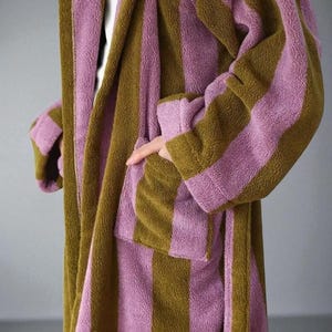 Op de afbeelding: Een gestreepte badjas in olijfgroen en lavendel. De badjas heeft brede verticale strepen, een sjaalkraag en zakken. De zachte, pluche stof is zichtbaar in de close-up. De badjas is een comfortabel kledingstuk om in te loungen.