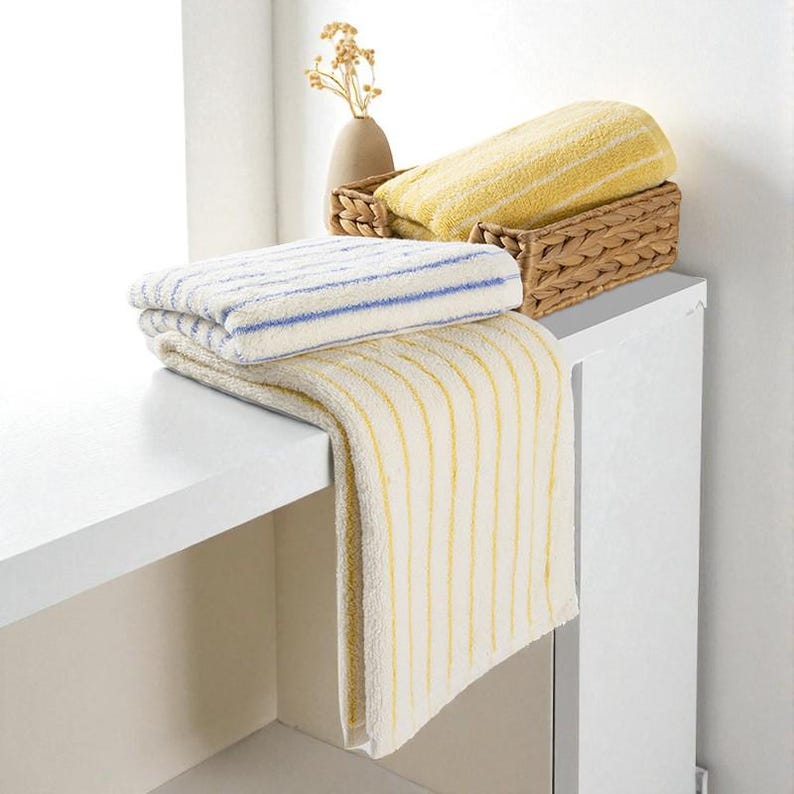 May include: A display of folded and draped bath towels in shades of yellow and cream, with blue and yellow stripes. One towel is draped over a white surface, while others are stacked or in a woven basket. A small vase with dried flowers is in the background.