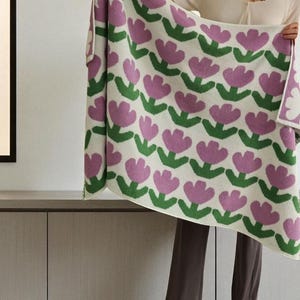May include: A cream-colored blanket with a repeating pattern of pink tulip flowers and green leaves. The blanket is held up, showcasing the floral design. The soft blanket is a cozy home decor item.