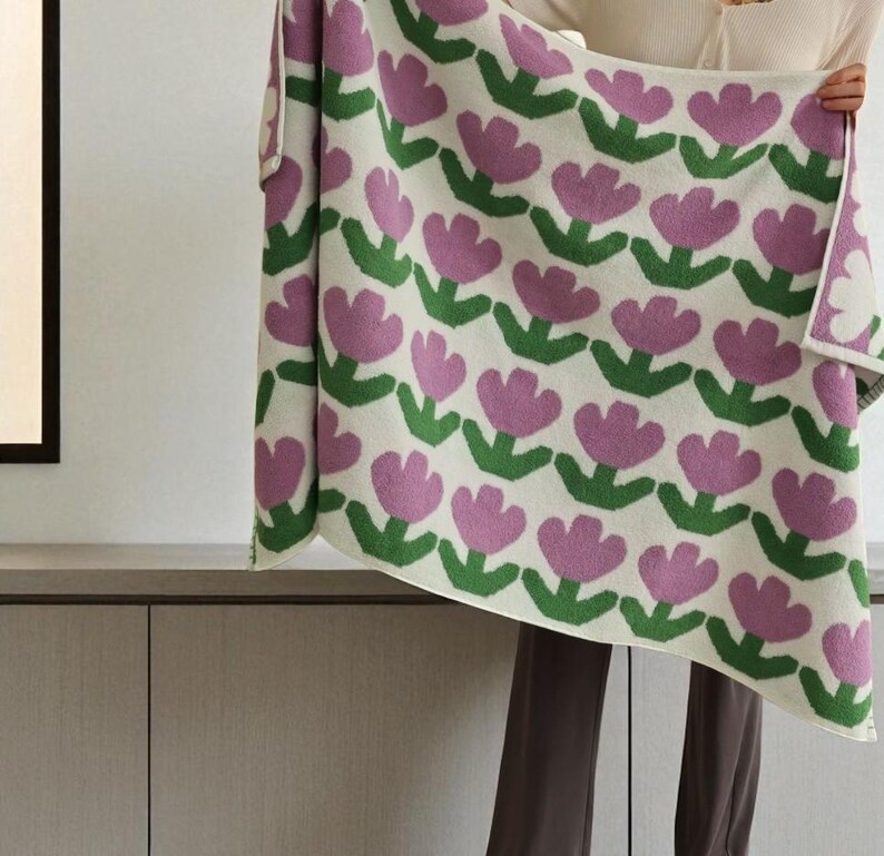 May include: A cream-colored blanket with a repeating pattern of pink tulip flowers and green leaves. The blanket is held up, showcasing the floral design. The soft blanket is a cozy home decor item.