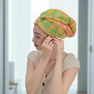 May include: A yellow hair towel with a pear pattern in green and pink. The towel is wrapped around the head and secured with a button. The person is wearing a light beige short-sleeved shirt.