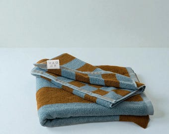 Op de afbeelding: Gevouwen blauw en bruin gestreepte handdoek. De handdoek heeft een dambordpatroon en een etiket met wasvoorschriften. De handdoek is gemaakt van een zacht, absorberend materiaal, perfect voor in de badkamer of op het strand.