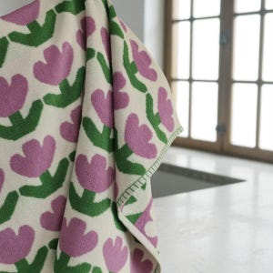 May include: A folded white towel with a repeating pattern of purple tulip-like flowers and green stems. The towel has a green stitched border and is hanging in front of a window.