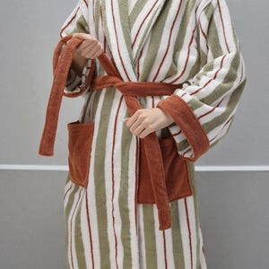 May include: A striped terry cloth robe in white, olive green, and rust red. The robe features a shawl collar, patch pockets, and a matching belt. The cuffs and pockets are trimmed in rust-colored fabric.