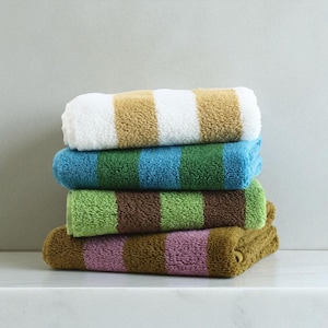 Op de afbeelding: Een stapel van vier opgerolde handdoeken met strepen in verschillende kleuren. De bovenste handdoek is wit en beige, gevolgd door blauw en groen, dan groen en bruin, en ten slotte bruin en roze. De handdoeken zijn op een wit oppervlak gestapeld.