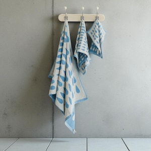 Könnte beinhalten: Drei passende Handtücher mit blau-weißem Muster hängen an einer weißen Stange vor einer grauen Wand. Die Handtücher weisen ein sich wiederholendes Tropfendesign auf. Die Handtücher sind unterschiedlich groß, wobei das größte Handtuch links hängt.