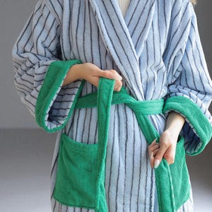 May include: A light blue and gray striped terry cloth robe with green trim and pockets. The robe has a matching green belt and long sleeves. The robe is made of a soft, absorbent material.
