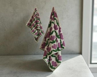 May include: Two off-white floral towels with a repeating pattern of purple flowers and green leaves. The towels are hanging against a gray wall. The edges of the towels are trimmed with green stitching.