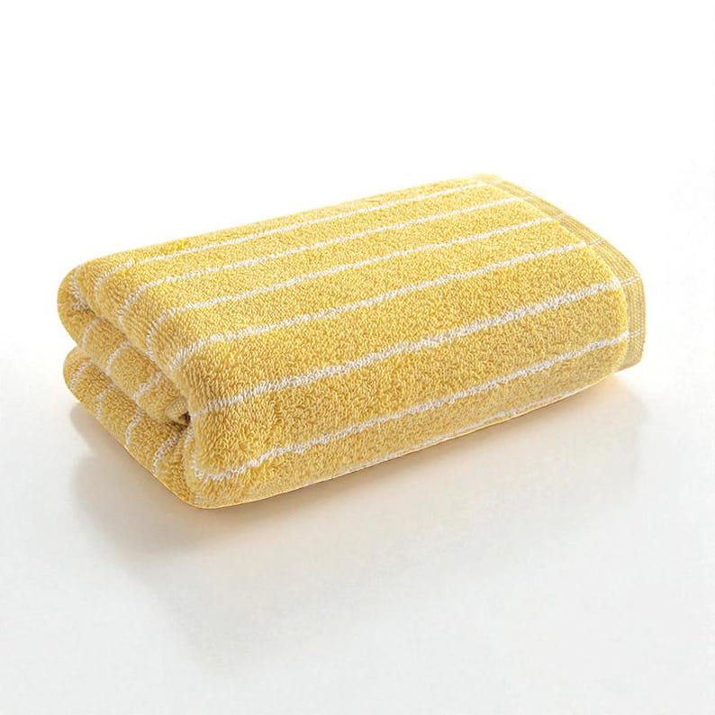 May include: Folded yellow bath towel with white horizontal stripes. The towel is made of a soft, absorbent material, perfect for use in a bathroom or spa setting. The bright color and simple design make it a versatile addition to any decor.