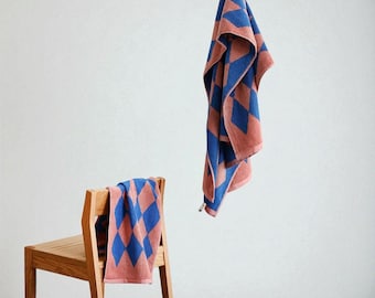 May include: Two bath towels with a blue and peach diamond pattern. One towel is draped over a wooden chair, and the other is hanging from a metal bar. The towels are made of a soft, absorbent material.