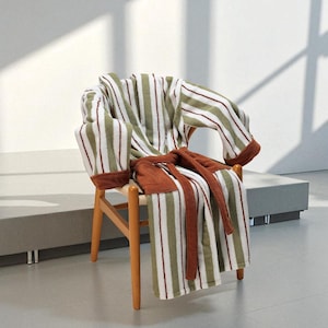 May include: A striped bathrobe in white, olive green, and burgundy rests on a wooden chair. The robe's belt is a solid brown, and the chair has a light wood frame. The scene is lit by natural light, creating shadows.