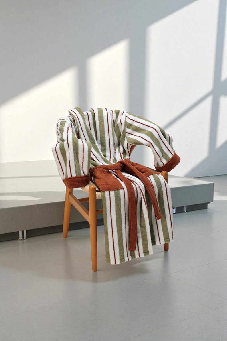 May include: A striped bathrobe in white, olive green, and burgundy rests on a wooden chair. The robe's belt is a solid brown, and the chair has a light wood frame. The scene is lit by natural light, creating shadows.