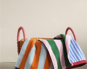 Op de afbeelding: Een gestreepte strandhanddoek in een rieten mand. De handdoek heeft oranje, lichtblauwe, groene, roze en blauwe strepen. De mand heeft roze handvatten en een groene rand. De handdoek is over de rand van de mand gedrapeerd.