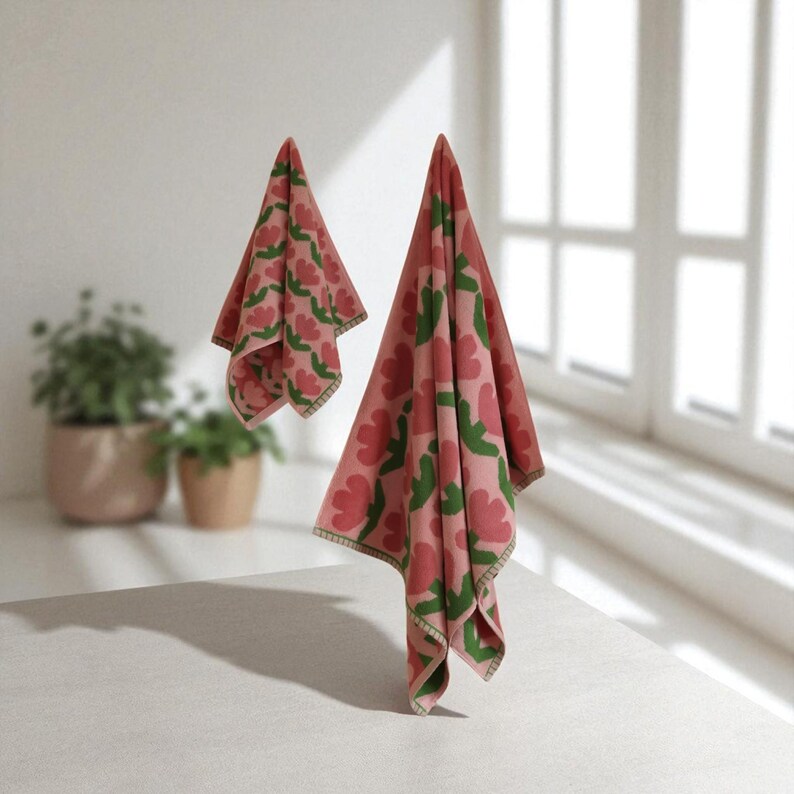 May include: Two pink and green floral patterned towels hanging in front of a window. The towels have a repeating pattern of red flowers and green leaves. The towels are made of a soft material and have a decorative border.
