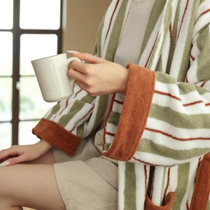 May include: A striped green and white robe with rust-colored accents. The robe has a shawl collar, pockets, and a belt. The person is holding a white mug with a brown rim. The robe's sleeves have rust-colored cuffs.