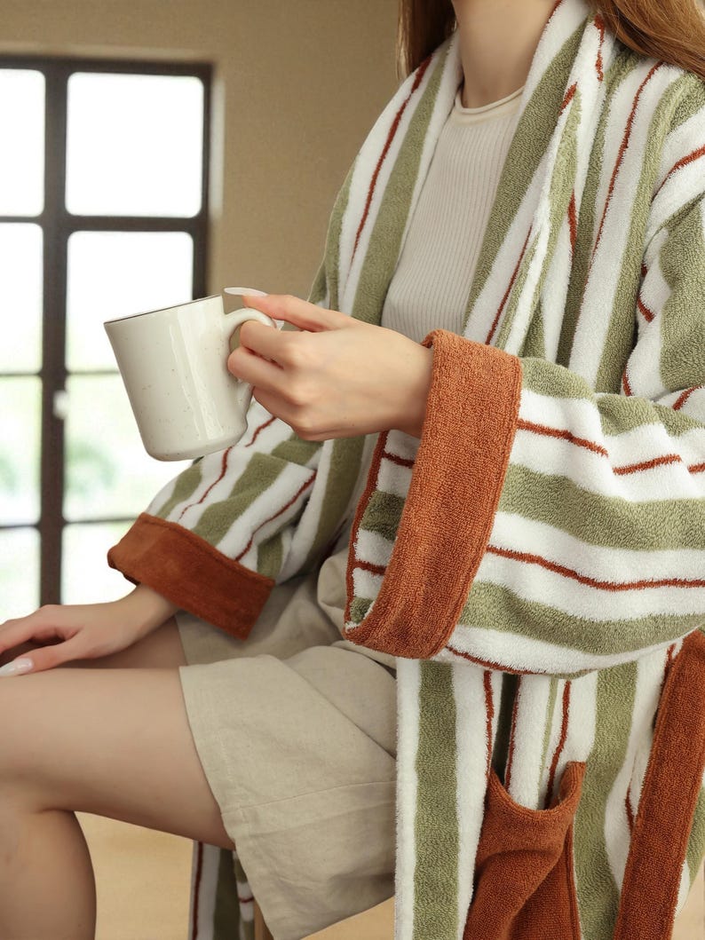 May include: A striped green and white robe with rust-colored accents. The robe has a shawl collar, pockets, and a belt. The person is holding a white mug with a brown rim. The robe's sleeves have rust-colored cuffs.