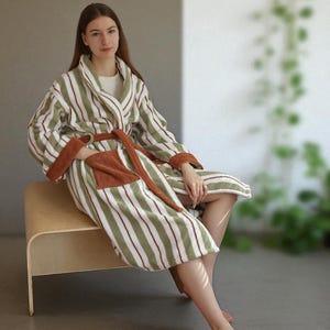 May include: A striped robe in olive green, white, and burgundy. The robe features a shawl collar, a belt, and pockets with a brown trim. The person is sitting on a wooden bench. The robe is made of a soft, plush material.