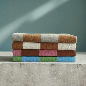 May include: A stack of four folded bath towels. Each towel has a colour-block design with alternating stripes of brown, grey, pink, green, and blue. The towels are stacked on a concrete surface against a teal background.