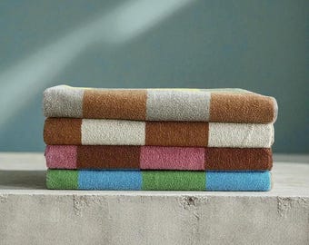 Op de afbeelding: Een stapel van vier opgevouwen badhanddoeken. Elke handdoek heeft een colorblock-ontwerp met afwisselende strepen in bruin, grijs, roze, groen en blauw. De handdoeken zijn gestapeld op een betonnen oppervlak tegen een turquoise achtergrond.