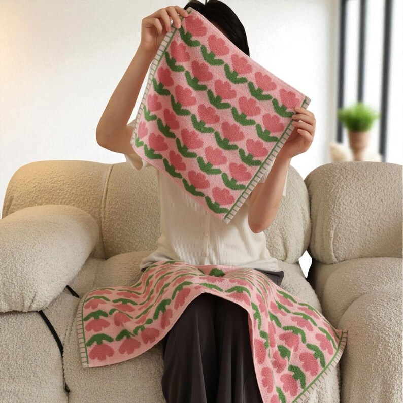 May include: Pink and green floral patterned towel. The towel features a repeating pattern of pink tulip-like flowers with green stems and leaves. The edges are trimmed with a green border. The towel is soft and appears to be made of a woven material.