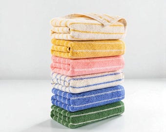 Op de afbeelding: Een stapel van zeven opgevouwen handdoeken in verschillende kleuren: crème met gele strepen, geel, roze, wit met blauwe strepen, blauw en groen. De handdoeken zijn netjes gerangschikt en tonen een reeks tinten en gestreepte patronen. De handdoeken zijn gemaakt van zacht, absorberend materiaal.