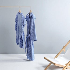 May include: Two blue and white striped robes hanging on a wooden rack. One robe is a short-sleeved, button-up style, and the other is a long robe with a hood and tie. A wooden and white canvas deck chair is in the background.