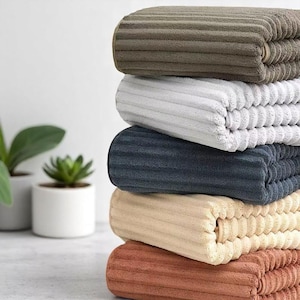 Puede incluir: Una pila de seis toallas de pana dobladas en varios colores: marrón, gris claro, azul marino, beige y óxido. Las toallas tienen una textura acanalada y están cuidadosamente apiladas. Las toallas son suaves y afelpadas.