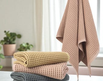 May include: A stack of four folded waffle-weave towels in shades of tan, pink, and gray. One matching towel hangs in the background. The towels are made of a textured material and are neatly arranged on a light-colored surface.