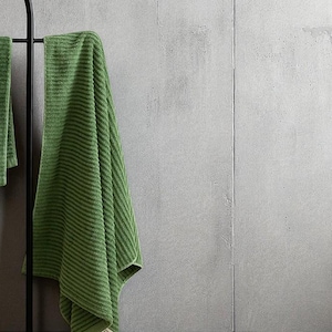 May include: Green bath towels with a ribbed texture hang on a black metal rack. The towels are a solid green colour and appear soft. The background is a light grey wall.