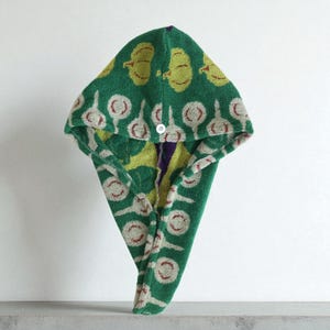 May include: A green hooded hair towel with a button closure. The towel features a repeating pattern of yellow apples and white and red circular designs. The towel is made of a soft, absorbent material.