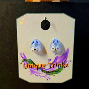 May include: A pair of light blue stud earrings shaped like cartoon animals with blue accents, displayed on a white card with the text "Unique Trinkx" in orange and a purple and green splash design.