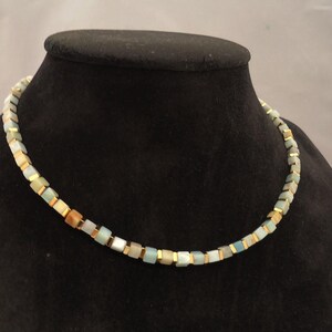 Può includere: Una collana di piccole perline quadrate nei toni del blu, verde, beige e oro. La collana è esposta su un busto di manichino nero. Le perline sono infilate con distanziatori color oro.