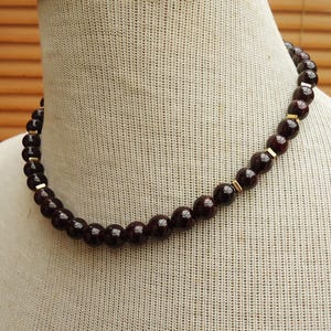 May include: A beaded necklace featuring dark red, round beads. The necklace is accented with small gold-colored rectangular spacers. The necklace is displayed on a neutral-colored mannequin.