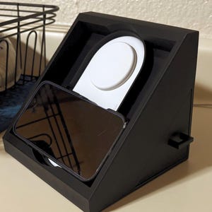 Może przedstawiać: Czarna stacja ładująca z białą okrągłą podkładką ładującą i czarnym smartfonem. Stacja jest ustawiona pod kątem i przeznaczona do ładowania telefonu i zegarka. Stacja znajduje się na jasnobeżowej powierzchni.