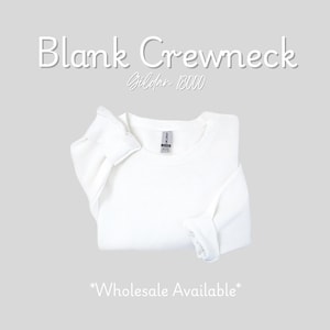 K&ouml;nnte beinhalten: Ein gefaltetes wei&szlig;es Crewneck-Sweatshirt mit dem Text "Blank Crewneck Gildan 18000" in Wei&szlig;. Die Worte "Wholesale Available" stehen am unteren Rand des Bildes. Das Sweatshirt befindet sich auf einem hellgrauen Hintergrund.