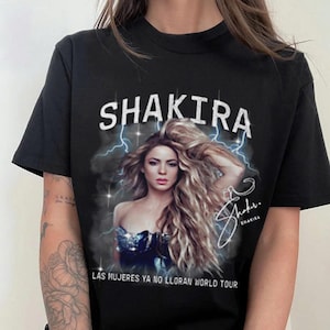 May include: Black t-shirt featuring the name "SHAKIRA" in white block letters above a portrait of the singer. The image includes her signature and the text "LAS MUJERES YA NO LLORAN WORLD TOUR".