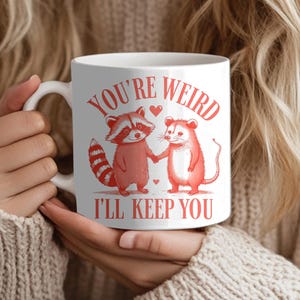 May include: White ceramic mug with the text "YOU'RE WEIRD I'LL KEEP YOU" in red. The mug features a cartoon raccoon and mouse holding hands. The raccoon has a striped tail and the mouse has a long tail.