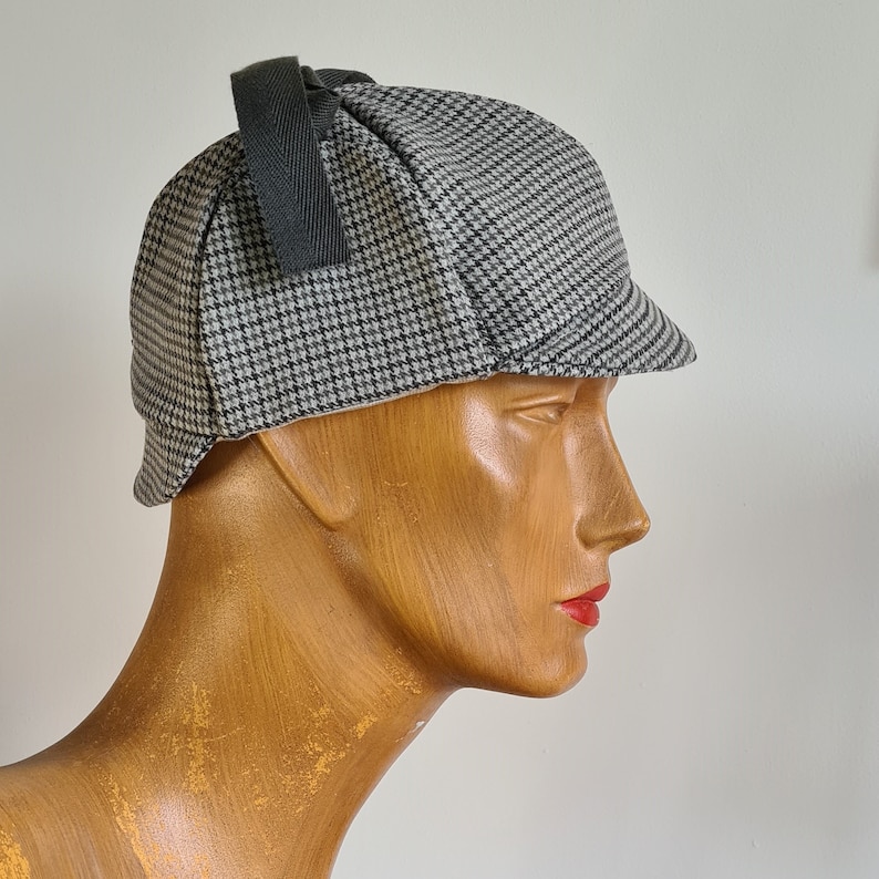 May include: A gray and black tweed hat with a black ribbon. The hat is worn on a wooden mannequin head.